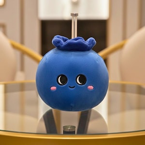 Könnte beinhalten: Ein blaues Plüschspielzeug in Form einer lächelnden Blaubeere mit schwarzen Augen und rosa Wangen. Die Blaubeere hat einen kleinen grünen Stiel und steht auf einem Glastisch. Das Design erinnert an eine Zeichentrickfigur.