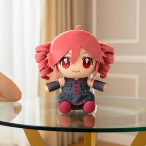 May include: A plush doll with a cartoon-like design. The doll has red hair styled in two large curls, red eyes, and a smiling expression. It wears a dark gray dress with red accents and matching red shoes. The doll is sitting on a glass table.