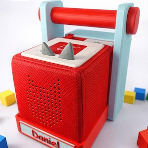 May include: A red, square audio player with a light blue handle and accents. The player features a speaker grill, a white top with grey shark fin-shaped buttons, and the name "Daniel". Colourful blocks surround the player.