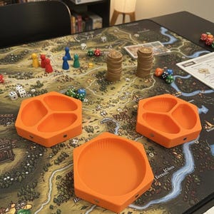 Könnte beinhalten: Draufsicht auf ein Brettspiel. Das Spielbrett zeigt eine Karte mit Spielfiguren, Würfeln, Holzmarkern und orangefarbenen Tabletts. Die Tabletts sind sechseckig und in Fächer unterteilt.