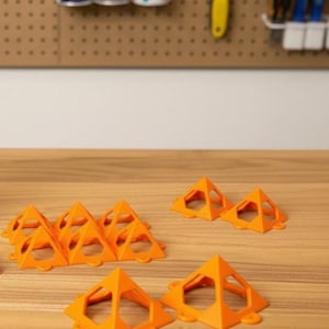 May include: Bright orange pyramid-shaped objects with a hollow center and rounded base are arranged on a wooden surface. The objects are likely used for a specific purpose, possibly in a craft or construction project. The background includes tools and supplies.