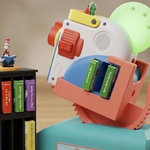 Op de afbeelding: Een 3D-geprinte speelgoedprojector met een witte behuizing, rode accenten en een lichtgroene lens, inclusief een kleine boekenplank met miniatuur Dr. Seuss-boeken en een Cat in the Hat-figuur. De basis is blauwgroen met de naam Ember erop.