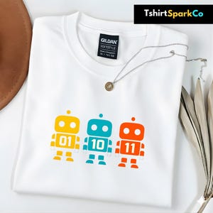 Peut inclure: T-shirt blanc avec un motif de robots colorés. Les robots sont jaunes, turquoise et orange, avec les chiffres 01, 10 et 11. Un collier argenté avec un petit pendentif est posé sur le t-shirt. Le nom de la marque "TshirtSparkCo" est visible.