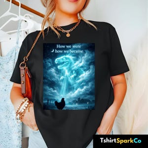 Puede incluir: Camiseta negra con un gráfico de un Tyrannosaurus Rex azul brillante emergiendo de las nubes, con una gallina debajo. El texto "How we were, how we became" está encima del dinosaurio. La camiseta tiene cuello redondo.