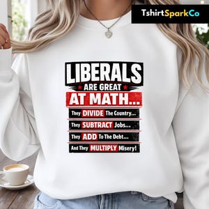 May include: White sweatshirt with the text "LIBERALS ARE GREAT AT MATH..." in black and red. The text continues with statements about dividing the country, subtracting jobs, adding to the debt, and multiplying misery. The brand name "TshirtSparkCo" is in the top right corner.