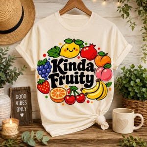 Może przedstawiać: Kremowa koszulka z kolorowym wzorem "Kinda Fruity" z różnymi owocami, takimi jak uśmiechnięta cytryna, winogrona, truskawki i wiśnie. Koszulka jest zawiązana na dole. Widoczny jest kubek z motywem serca.