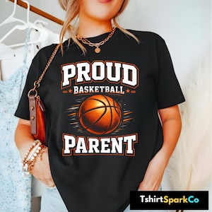Puede incluir: Camiseta negra con las palabras "PROUD BASKETBALL PARENT" en letras blancas y naranjas. Un gráfico de baloncesto está centrado debajo de la palabra "BASKETBALL". La camiseta tiene mangas cortas y cuello redondo.