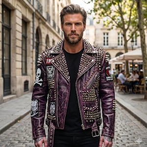 Op de afbeelding: Een verweerde paarse leren motorjas met zilverkleurige metalen spikes en studs op de revers, schouders en borst. De jas is voorzien van meerdere zwarte patches met witte grafische ontwerpen, waaronder een schedel met gekruiste beenderen.