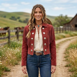 May include: A rust-colored suede jacket with fringe detailing and large silver buttons. The jacket is worn over a cream-colored blouse and paired with blue jeans. A brown leather belt and turquoise necklace complete the Western-inspired look.