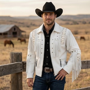 Pode incluir: Casaco de couro branco com franjas e tachas prateadas, usado com uma camisa preta, jeans azuis e um chapéu de cowboy preto. O casaco tem franjas nas mangas e nos ombros.