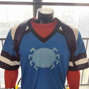 May include: A blue t-shirt featuring a light blue gear design. The shirt has black and white striped sleeves and a black V-neck collar. A black backpack is worn over the shirt.