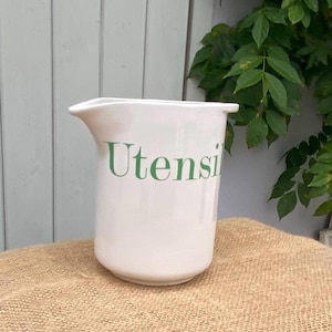 May include: A white ceramic utensil holder with a cylindrical body and a distinct pouring spout on one side. The word Utensils is printed in green serif lettering across the front of the smooth, glossy surface of the container.