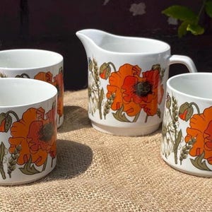 Puede incluir: Un juego de tres tazas de cerámica blanca y una jarra de leche a juego, decoradas con un vibrante patrón botánico que presenta grandes amapolas naranjas, tallos de trigo verdes y margaritas blancas sobre un fondo blanco.