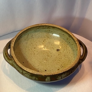 May include: A handmade ceramic bowl with two handles. The bowl has a speckled, light green interior and a darker green rim. The exterior has a textured, earthy appearance, suggesting a rustic or artisanal style. The bowl is suitable for serving food.