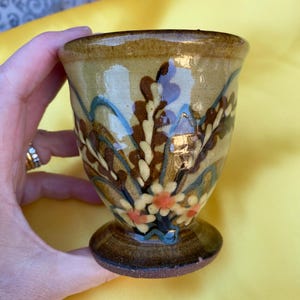 Puede incluir: Un cáliz de cerámica pintado a mano con base de pedestal. El cáliz presenta un fondo beige con diseños florales y de hojas en marrón, azul y naranja. El borde y la base son de color marrón oscuro.