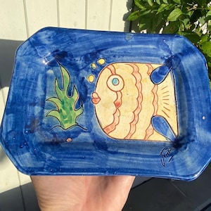 May include: A rectangular ceramic tray with a deep blue border and a central cream-coloured panel featuring a hand-painted orange fish with red stripes, a green aquatic plant, and yellow bubbles. The artist's signature is visible in the bottom corner.
