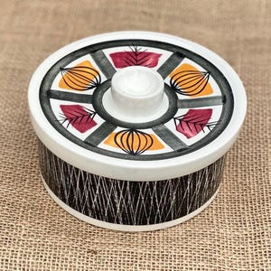 May include: A circular ceramic container with a lid. The lid has a white background with a black and grey geometric design and orange and maroon leaf-like shapes. The container's body has a black and white textured pattern.