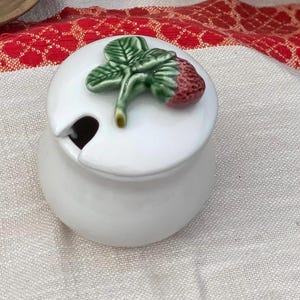 May include: A white ceramic jar with a rounded body and a matching lid. The lid features a sculpted, three-dimensional strawberry with green leaves and a red berry. A small notch is cut into the side of the lid to accommodate a spoon.