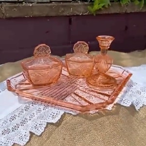 May include: A set of pink depression glass items including a square tray with a sunburst pattern, two lidded dishes with floral motifs, and a single candlestick holder. The glass pieces feature a translucent salmon-pink hue and molded details.