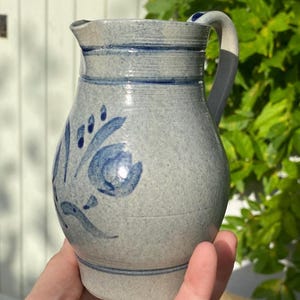 Pode incluir: Uma jarra de grés cinza-claro com textura salpicada e um motivo floral pintado à mão em azul. O recipiente apresenta uma alça curva, um bico vertedor e linhas horizontais incisas ao redor do pescoço e da base, com acabamento vidrado.