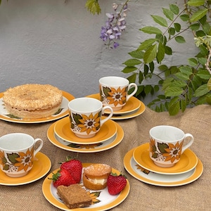 May include: A vintage tea set with yellow and white sunflower designs. The set includes teacups, saucers, and plates, arranged with cake, strawberries, and cookies. The scene is set on a burlap surface, with greenery in the background.