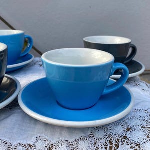 May include: A set of ceramic cups and saucers in bright blue and dark gray colors. Each cup features a smooth, rounded handle and a white interior. The saucers have a matching white rim, providing a clean contrast to the solid colored exterior.