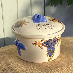 May include: A cream-coloured ceramic lidded container featuring blue and yellow decorative scrollwork patterns. The lid has a blue handle and the text "Poubelle de table" written in black script. Two blue moulded handles are on the sides.