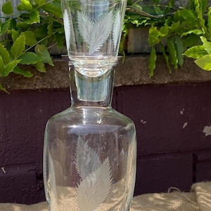 May include: A clear glass carafe with a matching glass tumbler resting on top as a lid. Both pieces feature an etched leaf pattern with fine serrated edges. The carafe has a rounded body and a narrow neck, while the tumbler is tapered.