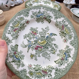 Pode incluir: Prato de cerâmica branco com um desenho floral e de pássaros em verde, azul, amarelo e rosa. O prato tem uma borda ondulada com uma borda verde a condizer. Outros pratos e chávenas são visíveis no fundo. Estilo vintage.