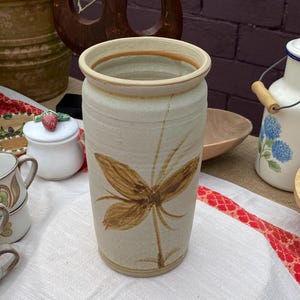 Op de afbeelding: Een beige keramische vaas met een bruin bloemmotief. De vaas heeft een brede opening en een licht gestructureerd oppervlak. Het ontwerp is een gestileerde bloem. De vaas staat op een wit tafelkleed.