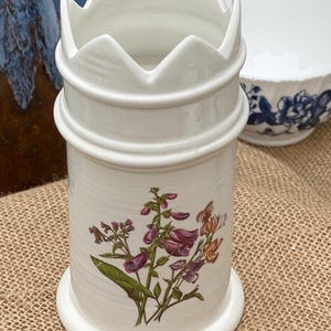 Op de afbeelding: Witte keramische keukengerei houder met een bloemenontwerp met paarse en oranje bloemen. De houder heeft een decoratieve kroonachtige bovenkant en een cilindrisch lichaam. Een witte kom met blauwe bloempatronen staat op de achtergrond.