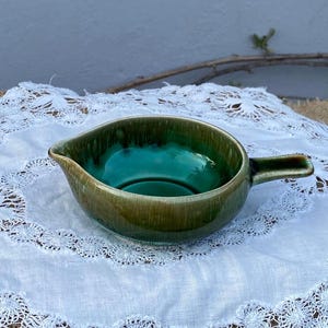 Puede incluir: Una salsera de cerámica con asa y pico vertedor. El interior es de un vibrante verde azulado, que contrasta con el esmalte verde y marrón terroso del exterior. La salsera está colocada sobre un mantel de encaje blanco.