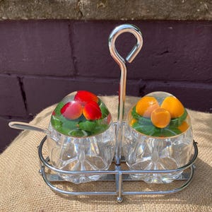 Peut inclure: Un ensemble de condiments composé de deux pots en verre transparent à base facettée, maintenus dans un support en métal chromé avec une poignée centrale. Chaque couvercle transparent contient des fraises rouges ou des tranches d'agrumes orange.