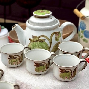 Peut inclure: Service à thé en céramique blanche comprenant une théière et plusieurs tasses. La théière a un couvercle et une anse, et les tasses ont des anses. Chaque pièce est décorée d'un motif floral vert et marron. L'ensemble est sur une surface blanche.