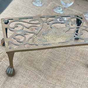 May include: A vintage brass trivet featuring a swan motif. The trivet is rectangular, with decorative cut-out patterns and four claw feet. The swan is centrally positioned. The trivet measures approximately 18 cm long and 13 cm wide.