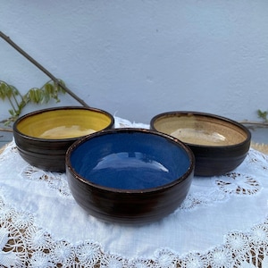 May include: Three ceramic bowls with a dark brown exterior and colorful interiors. One bowl has a yellow interior, another has a blue interior, and the third has a tan interior. The bowls are arranged on a white lace cloth.
