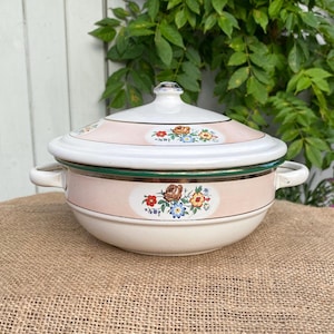 May include: A white enamel casserole dish with a lid, featuring a pale pink horizontal band around the centre and lid. The dish is decorated with a floral motif of red, brown, yellow, and blue flowers, accented by a thin metallic silver rim.