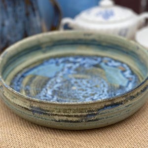 May include: A shallow, round ceramic bowl with a textured, mottled exterior in shades of muted green and brown. The interior features a deep blue glaze with a swirling, intricate pattern that transitions into lighter blue and cream tones at the center.