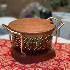 Puede incluir: Un tarro de cerámica marrón con tapa de madera y marco de alambre metálico. El tarro presenta un patrón de tejido de cesta texturizado y el texto en relieve APPLE SAUCE. Se asienta sobre una base de madera y la tapa incluye una muesca.