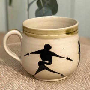 May include: Cream ceramic mug featuring a black silhouette of a person in action. The mug has a rounded shape, a handle, and a green band around the rim. The mug is approximately 10 cm tall.
