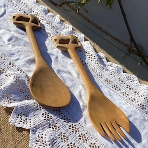Puede incluir: Dos utensilios de ensalada de madera, una cuchara y un tenedor, se exhiben sobre un mantel de encaje blanco. Los mangos tienen un diseño decorativo recortado. La madera tiene un color marrón claro natural.