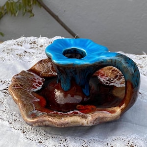 May include: A ceramic candle holder featuring a wide, wavy-edged base with a glossy brown and cream glaze. A central, bright blue flower-shaped cup rises from the centre, connected to the base by a curved handle with a matching blue glaze.