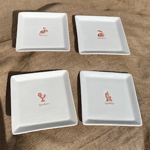 May include: Four white square ceramic appetizer plates. Each plate features a small, centred illustration in a reddish-brown tone: a coiled snack, two cherries, a cocktail glass with a citrus slice, and a bottle with a glass. Each has the word Appetizer.