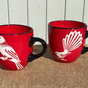 May include: Two bright red ceramic mugs with black handles and rims. Each mug features a white graphic illustration of a bird. The left mug displays a bird perched on a branch, while the right mug shows a bird with a large, fanned-out tail.