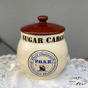 Puede incluir: Azucarero de cerámica color crema con tapa marrón. El tarro tiene las palabras "SUGAR CARGO" en negro y un gráfico circular azul y blanco con un barco y las letras "P.O.S.H."