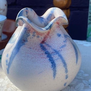 May include: A ceramic vase with a ruffled rim. The vase is mainly white with blue and red abstract designs. The vase has a rounded shape and sits on a patterned surface. The vase is likely used for decorative purposes.