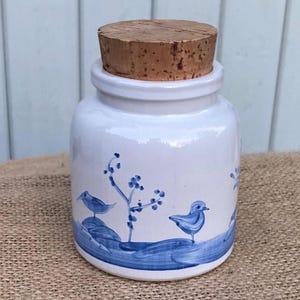 Könnte beinhalten: Ein weißes Keramik-Vorratsglas mit zylindrischem Körper und einem Naturkorken. Die Außenseite ist mit handgemalten blauen Illustrationen von kleinen Vögeln verziert, die auf Hügeln neben dünnen Zweigen sitzen.