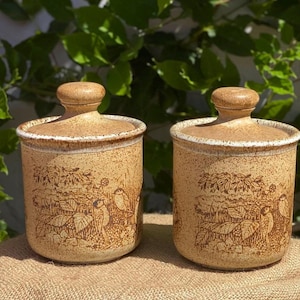 May include: Two matching ceramic canisters with lids. The beige-coloured canisters have a speckled texture and a brown illustration of birds and foliage. The lids are round with a small knob on top.