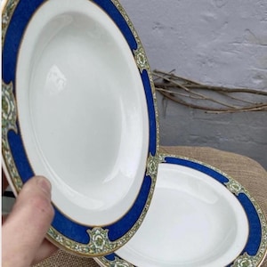 May include: Two white oval serving platters with a blue border and gold trim. The blue border is accented with a repeating floral design in green and gold. The platters are on a burlap surface.