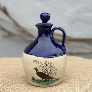 May include: A ceramic jug with a glossy dark blue upper section and a cream-colored base. The body features a hand-painted illustration of a pheasant standing near a small tree. The jug includes a matching dark blue lid with a round knob handle.